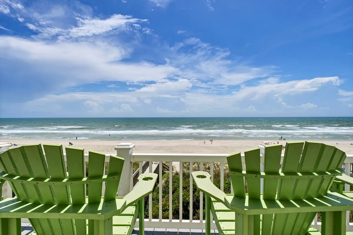 The hero image showcases a row of inviting green Adirondack chairs set on the deck of a spacious beachfront house, overlooking the endless sands of North Topsail Beach and the sparkling Atlantic Ocean under a sky dotted with soft clouds. The scene evokes tranquility, with the chairs perfectly positioned for sunrise views, relaxing, or gathering with loved ones to savor the gentle ocean breeze.
