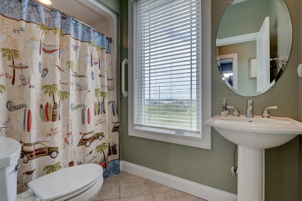 A colorful bathroom featuring a playful shower curtain, round mirror, and a window that lets in cheerful natural light. The decor is inviting, modern, and evokes a beach-inspired vibe.
