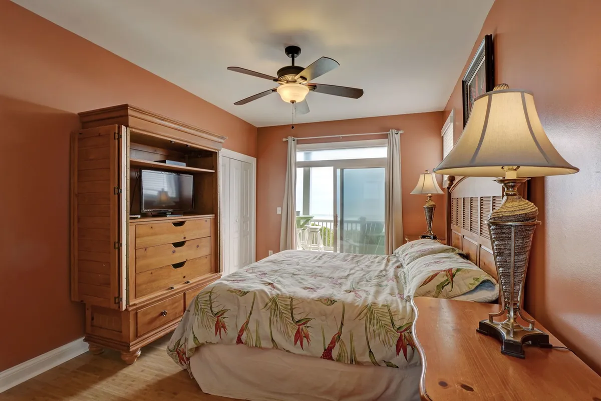 This image features a cozy bedroom with a bed dressed in floral bedding, a wooden dresser with a TV, and a large window offering a tranquil view. The room feels bright and inviting, ideal for personal comfort and relaxation.