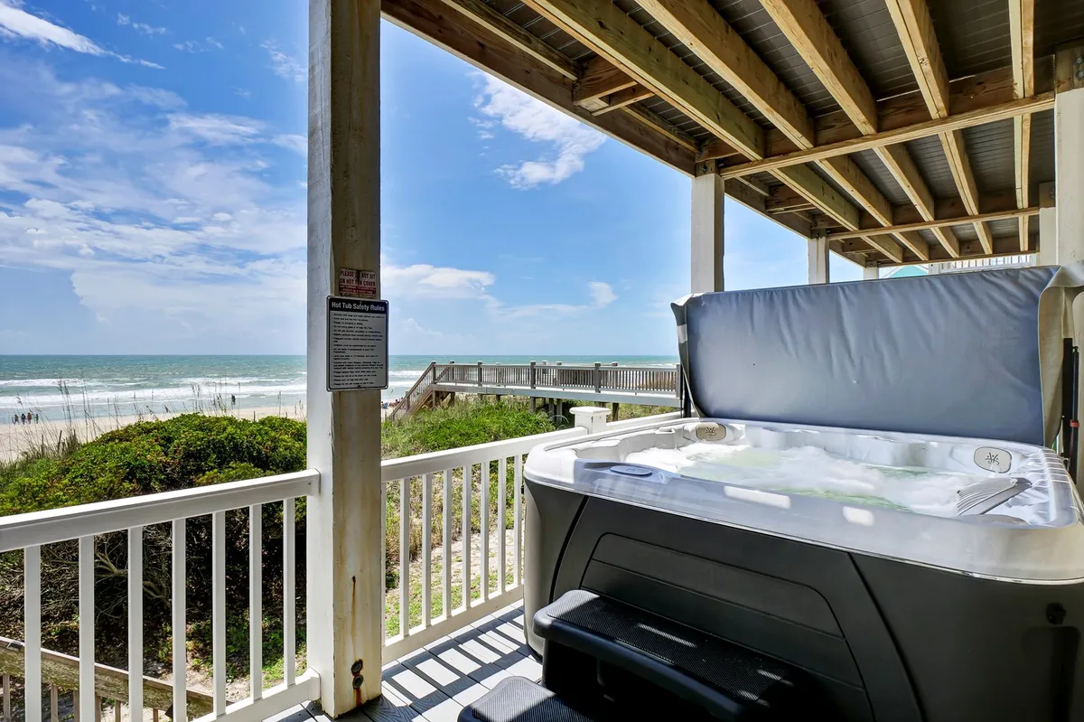 The image features a wooden deck directly overlooking the ocean, with a bubbling hot tub invitingly nestled under the deck area. The view showcases easy access to the sandy beach just steps away, with ample space for relaxation and socializing. The tranquil ambiance is heightened by the open horizon and gentle waves in the background, emphasizing privacy and luxury.