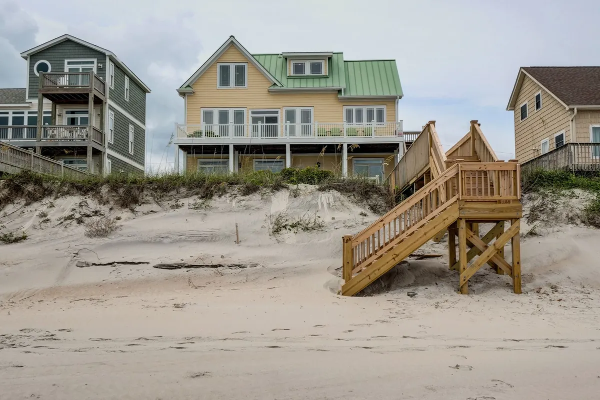 The image shows a beach house with a green metal roof, a wooden staircase leading directly to the sandy beach, and ample space for gatherings and outdoor fun. The inviting deck area emphasizes seamless indoor-outdoor living and access to the beach.