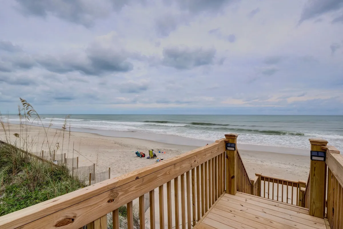 A spacious wooden deck overlooks both the beach and ocean, furnished with inviting chairs perfectly positioned for sunrise coffees or sunset watching. The proximity to the beach is clear, and the setting radiates relaxation and connection with nature.
