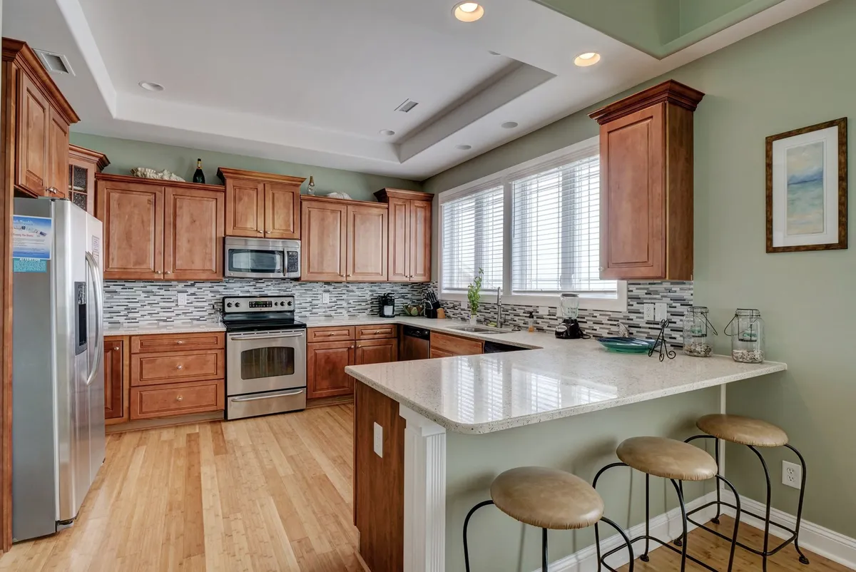 A bright kitchen space with a central island, stainless steel appliances, wood cabinetry, and large windows letting in ample sunlight. The setup invites cooking and entertaining, and hints at the potential for shared culinary adventures and family meals.