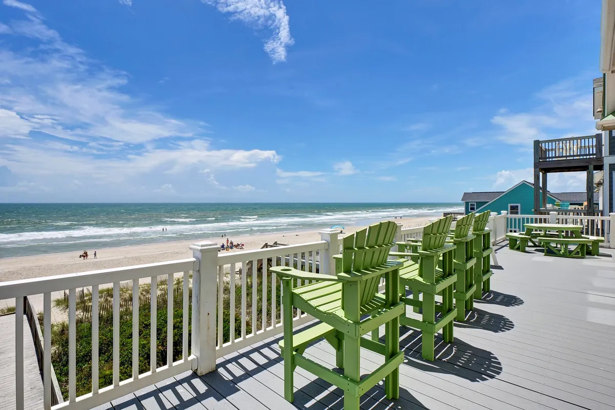 Green Adirondack chairs positioned on a deck overlook the broad expanse of the beach and ocean, with blue skies stretching above. The set-up invites guests to relax, socialize, and watch the sun set over the water.