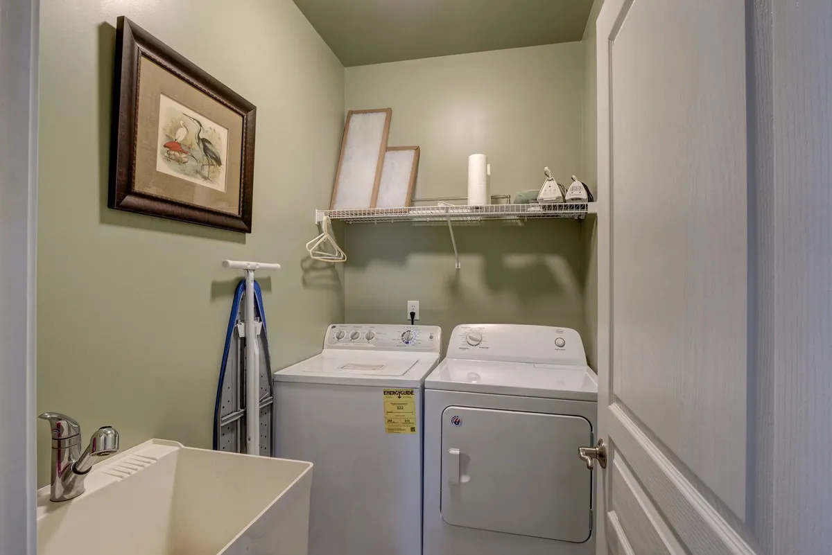 A practical laundry room with a washing machine, dryer, utility sink, framed artwork, and a shelf stocked with supplies. The area is neat and organized, emphasizing self-sufficiency and the ease of maintaining your own routines.