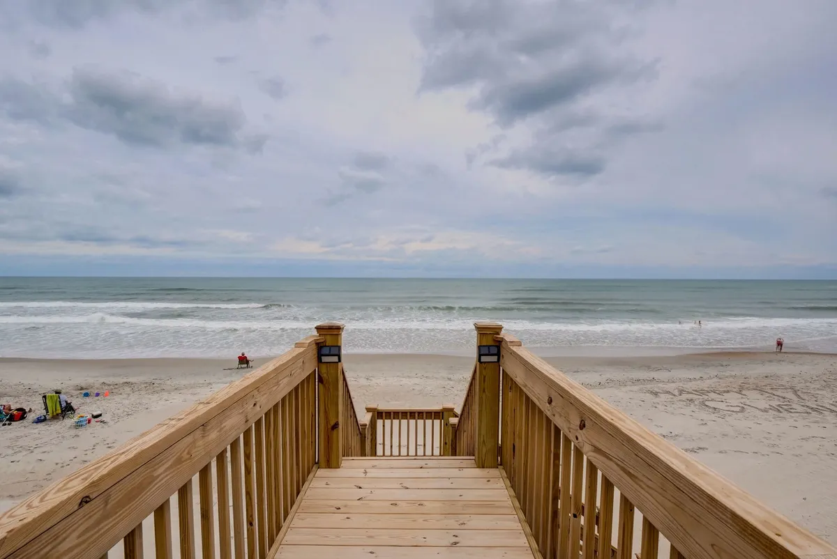 This image captures a wooden deck overlooking the ocean under a dramatic, cloudy sky. The expansive ocean view is front and center, suggesting a serene spot for morning coffee or evening relaxation while enjoying the sights and sounds of the waves.