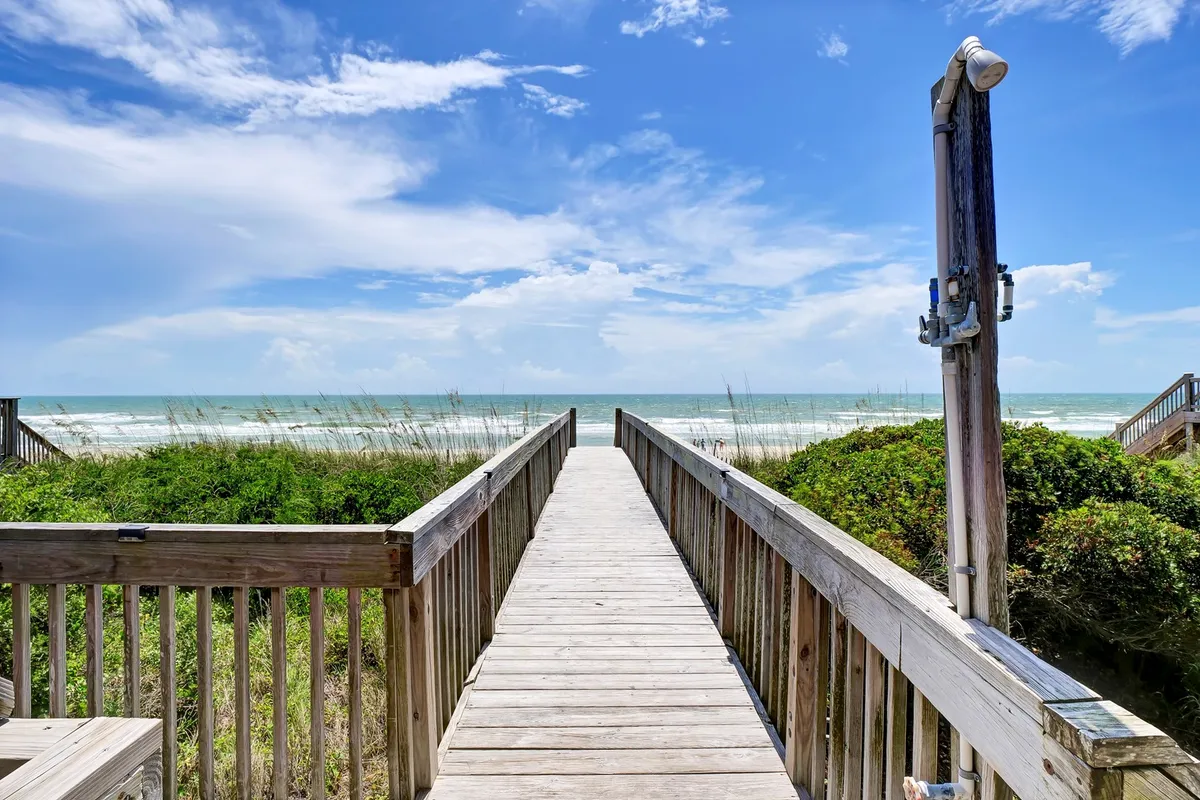 A wooden walkway winds through dune vegetation, leading straight to the open, sandy beach and sparkling ocean beyond. The perspective invites the viewer to imagine walking from the home directly onto the shore, surrounded by natural beauty and tranquility.