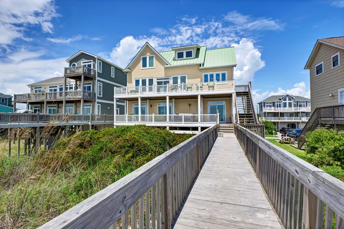 A large coastal house with multiple balconies and a wooden walkway leading to the entrance, surrounded by greenery. The home’s position and porches suggest close proximity to the water and outdoor gathering spaces.
