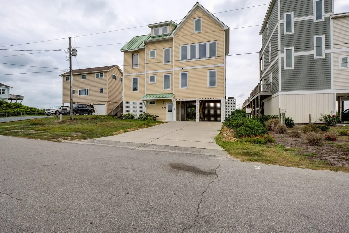A wide view of a three-story yellow beach house with a green metal roof, surrounded by other homes and a generous driveway. The multiple balconies and inviting architecture evoke a welcoming, resort-like atmosphere ideal for group stays.