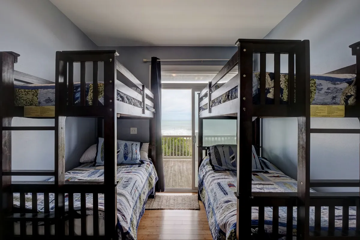 A cozy interior bunk room featuring bunk beds and an ocean view through the door leading to the deck. The space looks child-friendly and perfect for sleepovers or playtime after a day of beach activities.